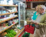 Starsza para z koszykiem i listą zakupów w sklepie spożywczym, obok półek z produktami.