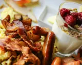 Herzhafte Ideen zum Brunch: Rührei, Speck, Würstchen und Kartoffeln neben einem Glas Joghurt mit Beeren und Müsli.