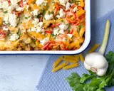 Herzhafter Gyros Auflauf mit Penne, Paprika und Feta, garniert mit Petersilie. Daneben liegen Penne, Knoblauch und frische Kräuter.