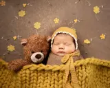 Bambino addormentato con orsacchiotto, avvolto in una coperta gialla. Un'idea per foto neonati fai da te con decorazioni floreali.