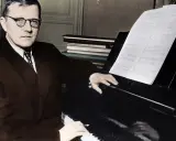 Dmitri Szostakowicz w okularach siedzi przy fortepianie, grając z nut.