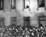 Menge jubelt vor Gebäude, während ein Mann auf einer Leiter steht. Die Szene erinnert an die Zeit des Ermächtigungsgesetzes.