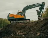 Pomarańczowo-czarna koparka Volvo na placu budowy, z innym mniejszym sprzętem w tle.