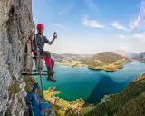 Mężczyzna w kasku siedzi na drewnianej platformie przy skalnej ścianie, ciesząc się widokiem na jezioro w Austrii. To wspaniała przygoda na via ferrata Austria!
