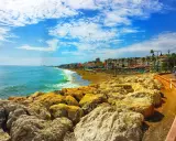 Sonniger Tag an der playa de la carihuela. Türkises Meer, goldener Sandstrand mit Liegen und Palmen, gesäumt von bunten Gebäuden. Felsen im Vordergrund.