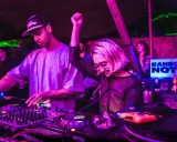 DJ set b2b: un uomo e una donna mixano musica in un locale con luci viola e verdi. La folla balla.