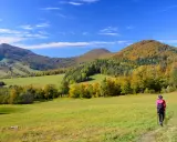 Jesień na **Beskid Niski szlaki**. Turystka w różowej kurtce idzie przez zieloną łąkę w kierunku kolorowych wzgórz.