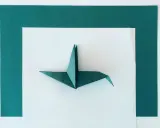 Zielony żuraw z papieru, przykład sztuki origami dla dorosłych, leży na białym tle otoczonym ciemnozielonymi ramkami.