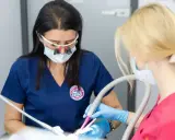 Dwie dentystki w maseczkach i okularach powiększających pracują przy pacjencie. Jedna używa wiertła, druga asystuje, podając anestetyki.