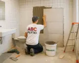 Fachowiec z koszulką "ABC PŁYTKI" układa płytkę na płytkę w łazience, używając kleju. Obok wiadro z zaprawą i drabina.