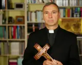Duchowny z krzyżem w dłoniach, na tle regałów z książkami. Czy religia wlicza się do czerwonego paska?