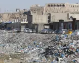 Müllberge säumen die Straßen von Kirkuk, Irak. Autos parken hinter den Geschäften, während im Hintergrund alte Gebäude auf einem Hügel stehen.