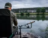 Wędkarz w kamuflażu siedzi na krześle nad jeziorem, gotowy do łowienia. Jego wędka feeder 3m spoczywa na podpórce, czekając na branie.
