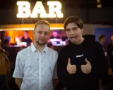 Dwóch mężczyzn pozuje przed napisem "BAR". Jeden w koszuli w palmy, drugi w bluzie z napisem SBM, pokazuje kciuki w górę i wystawia język.
