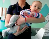 Dziecko w kolorowym ubranku, trzymane przez opiekunkę, przyjmuje pozycję amfibii. W tle widać elementy rehabilitacyjne.