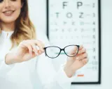 Okulary z astygmatyzmem – czy trzeba nosić cały czas? Optometrystka prezentuje okulary na tle tablicy Snellena.