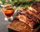 Costine al forno succulente e caramellate, servite su tagliere di legno con salsa barbecue in ciotolina.