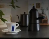 Eine Tasse Lavazza-Kaffee steht neben einer schwarzen Mokkakanne. Die Szene vermittelt die klassische italienische Kaffeezubereitung.