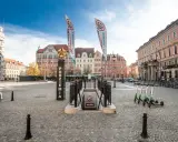 Wejście do podziemi z napisem "Movie Gate" i dwoma pionowymi banerami. W tle kolorowe kamienice i błękitne niebo.