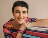 Uśmiechnięta kobieta z krótkimi włosami opiera się o stos kolorowych książek. Wiedza, którą posiadają, jest dla niej dostateczna.