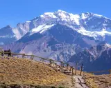 Widok na majestatyczne góry z ośnieżonym szczytem Aconcagua. Ścieżka z drewnianym ogrodzeniem prowadzi przez suchą, trawiastą przestrzeń.