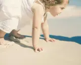 Małe dziecko w białej sukience raczkuje po plaży. Może to być jedna z przyczyn nie chodzenia dziecka, bo piasek jest ciekawy.