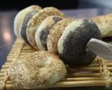 Une rangée de bagels faits maison, certains recouverts de graines de sésame, d'autres de pavot, sur une natte en bambou.