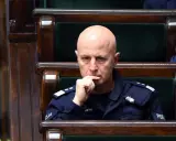 Łysy mężczyzna w mundurze policji siedzi w drewnianej ławie, zamyślony.