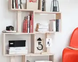 Bibliothèque DIY en bois clair, remplie de livres, d'une caméra vintage et d'une boîte noire. Une chaise rouge vif est visible à droite.