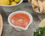 En skål med en röd, kryddig sås, kanske en pico de gallo, står på ett grått bord. Bredvid syns gula citroner och bröd.