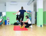 Kobieta ćwiczy propriocepcję na poduszce balansującej, druga osoba asekuruje.