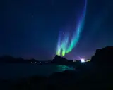 Gröna och lila norrsken dansar över en stjärnklar himmel i norra Sverige. En mörk silhuett av berg och en glittrande sjö syns nedanför.