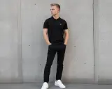Młody mężczyzna w czarnej koszulce polo i jeansach pokazuje, jak nosić koszulkę polo na co dzień.