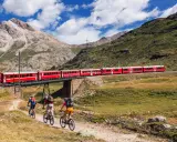 Treno rosso attraversa un ponte su un paesaggio alpino mozzafiato. Tre ciclisti pedalano su un sentiero sottostante, godendosi la vista della ferrovia del Bernina.