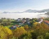 Pueblo en el **valle del Baztan** cubierto por niebla matutina, con casas blancas y tejados rojos entre árboles otoñales.