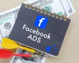 Notatnik z logo Facebook i napisem "Facebook ADS" leży na banknotach dolarowych. Obok znajdują się rzutki, symbolizujące celne reklamy mobilne.