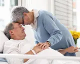 Czule pożegnanie. Kobieta pochyla się nad umierającym mężem, jak poznać że chory na alzheimera umiera, widać troskę i miłość.