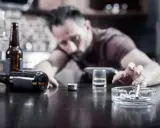 Mężczyzna z papierosem w popielniczce, butelki i kieliszek na stole. Nadmierne spożycie alkoholu może pogorszyć hemoroidy.