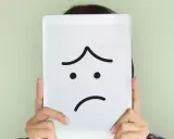 Una persona nasconde il volto dietro un tablet con un'espressione triste disegnata. La famiglia invischiata Minuchin potrebbe sentirsi così.