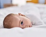 Bebé acostado sobre sábanas blancas, mirando hacia arriba con un ojo visible.