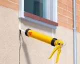 Pistola de calafateo amarilla aplicando sellador blanco para como tapar grietas en la pared exterior de estuco junto a una ventana.