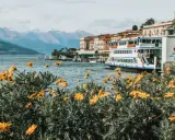 Fiori gialli in primo piano, lago blu, montagne innevate e un battello attraccato. Una perfetta gita fuori porta Milano.