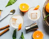 Zwei cremige Gin Sours mit Eis und Orangenzesten, garniert mit Zimtstangen und frischen Mandarinen.