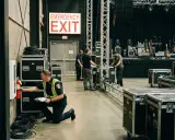 Pracownik sprawdza gaśnicę przy znaku "EMERGENCY EXIT". W tle widać scenę koncertową i sprzęt. To miejsce zbiórki do ewakuacji.