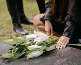 Dłoń kładzie białe lilie na kamieniu nagrobnym. W tle widać nogi w czarnych butach. To symboliczny gest, który może nastąpić w dowolnym momencie po śmierci, nie ma ustalonego terminu, ile dni po śmierci jest pogrzeb.