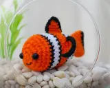 Adorable pez payaso de ganchillo en una pecera con piedras blancas y plantas verdes. ¡Perfecto para patrones de peces de ganchillo!