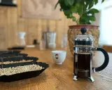 Eine French Press mit frisch gebrühtem Kaffee steht neben einer Tasse und Schalen mit Kaffeebohnen.