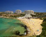 En livlig strand med turkost vatten och gyllene sand vid Golden Bay, Malta. Flera hotell syns i bakgrunden.