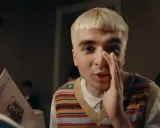 Młody mężczyzna z blond fryzurą, w kamizelce z kolorowym wzorem, trzyma rękę przy ustach, jakby coś szeptał. Obok niego leży blok mata tekst.