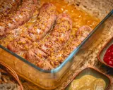 Pyszna biała kiełbasa w piwie, posypana ziarnami gorczycy, pieczona w szklanym naczyniu. Obok sosy i chleb.